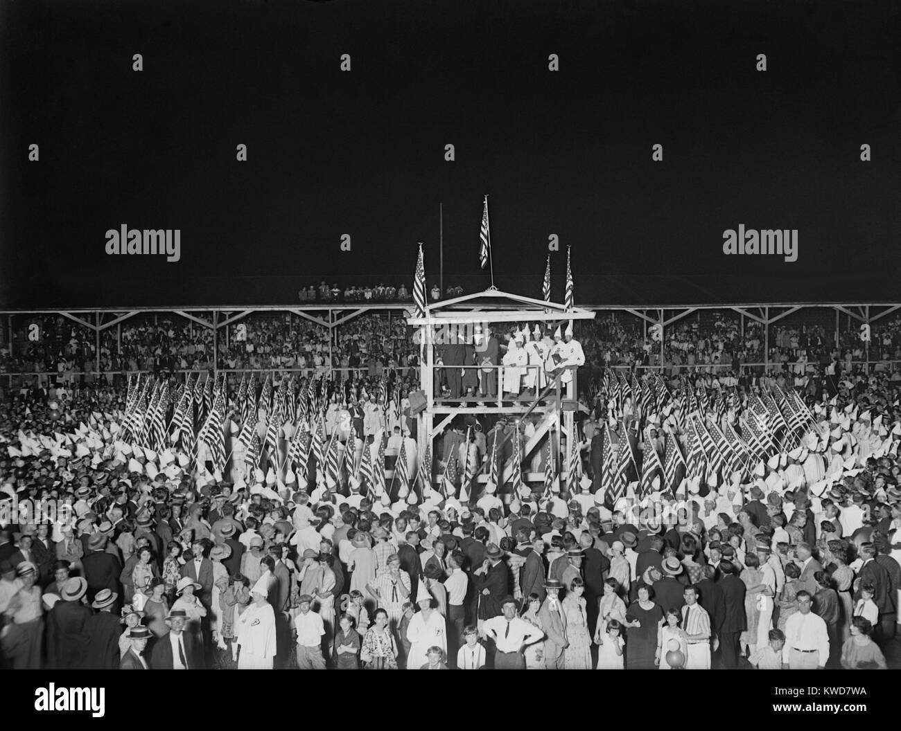 Ku Klux Klan nighttime ceremony during the great gathering in Washington, D.C. August 8, 1925. Klansmen hold dozens of American Flags in a circle around a raised Speaker's platform. Outside of the circle are a milling crowd of men, women, and some families. (BSLOC 2015 16 178) Stock Photo