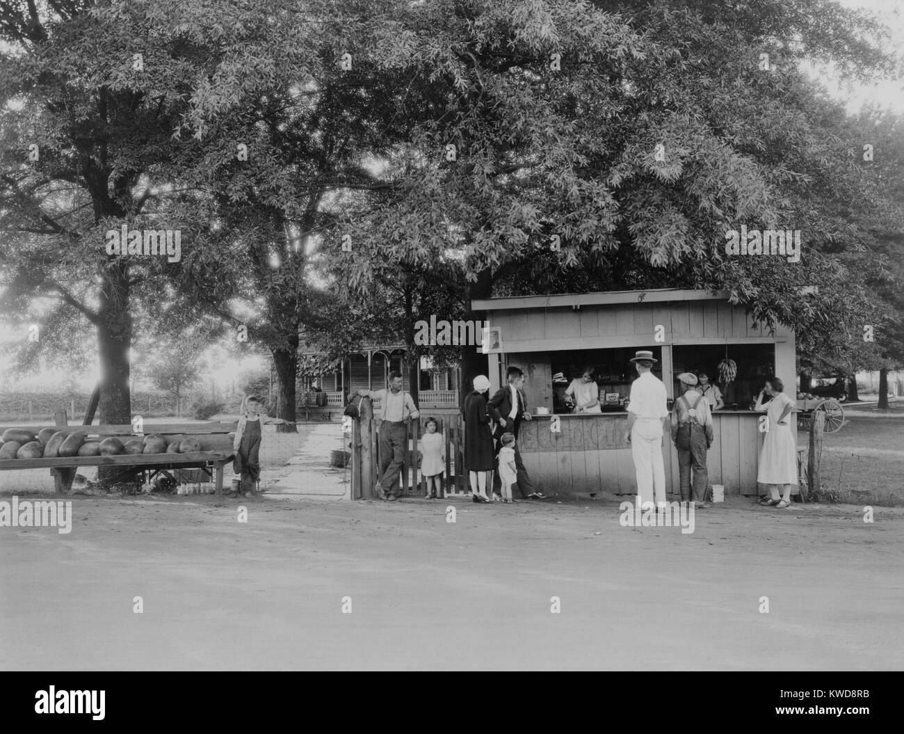 City folk in their high heels, cloche hat, suit, and neat toddler clothes at roadside food stand. 1925-30. The rural child is her barefoot, next to her father who wears heavy shoes and stained workpants (BSLOC 2016 8 110) Stock Photo