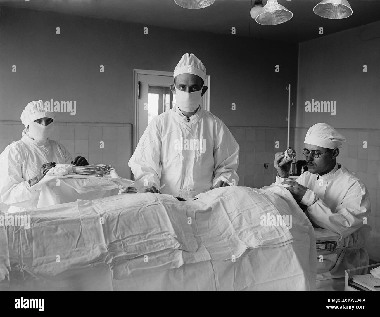 Surgical team before the operation begins, with patient is covered with sterile white cotton fabric. The anesthesiologist at right monitors patient's consciousness. 1922 in a hospital in Washington, D.C. (BSLOC_2016_10_34) Stock Photo