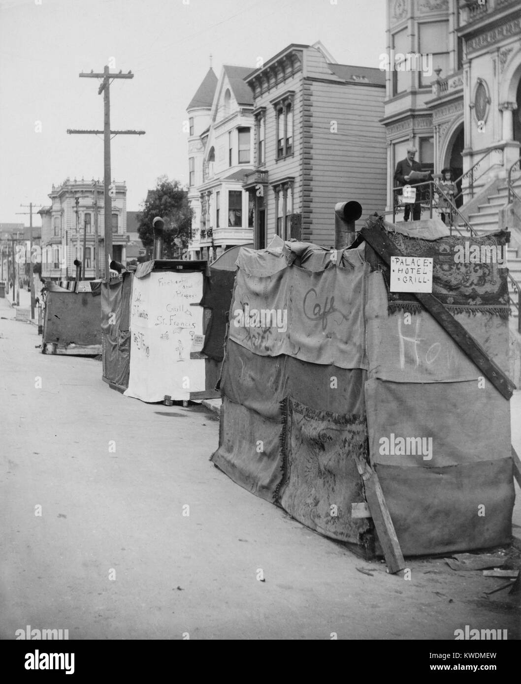 Temporary shelters in street after the April 18, 1906 San Francisco, earthquake and 3-day fire. A hand written sign identified the burlap and rug-covered hovel as the Palace Hotel Grill. A man stands reading a newspaper on his front steps of his intact home. People could live in their stable homes, but could not cook inside until houses were inspected for gas leaks that would case explosions and/or fires. (BSLOC 2017 17 47) Stock Photo