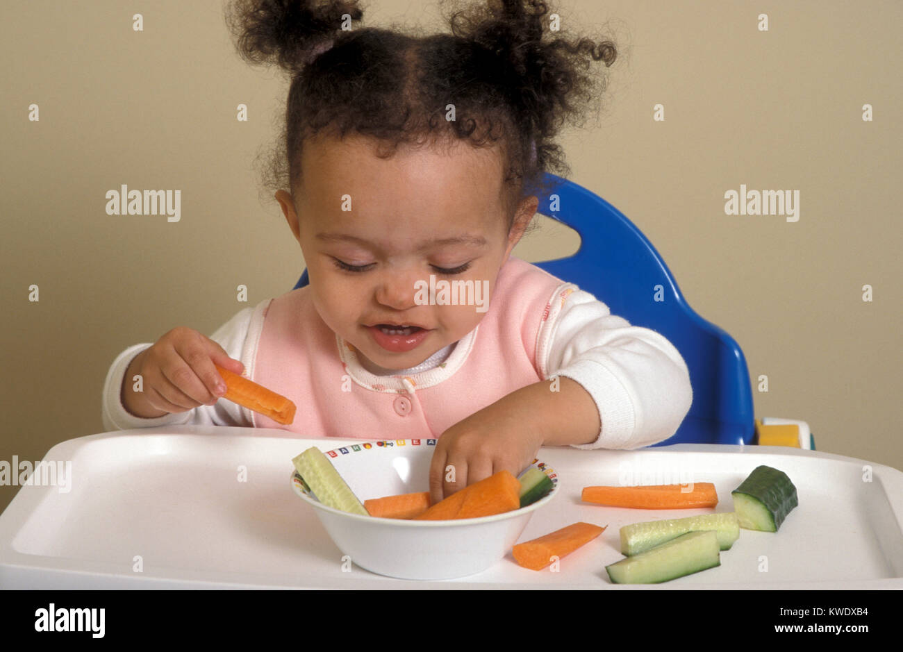mixed race baby in high chair eating finger foods of carrot and ...