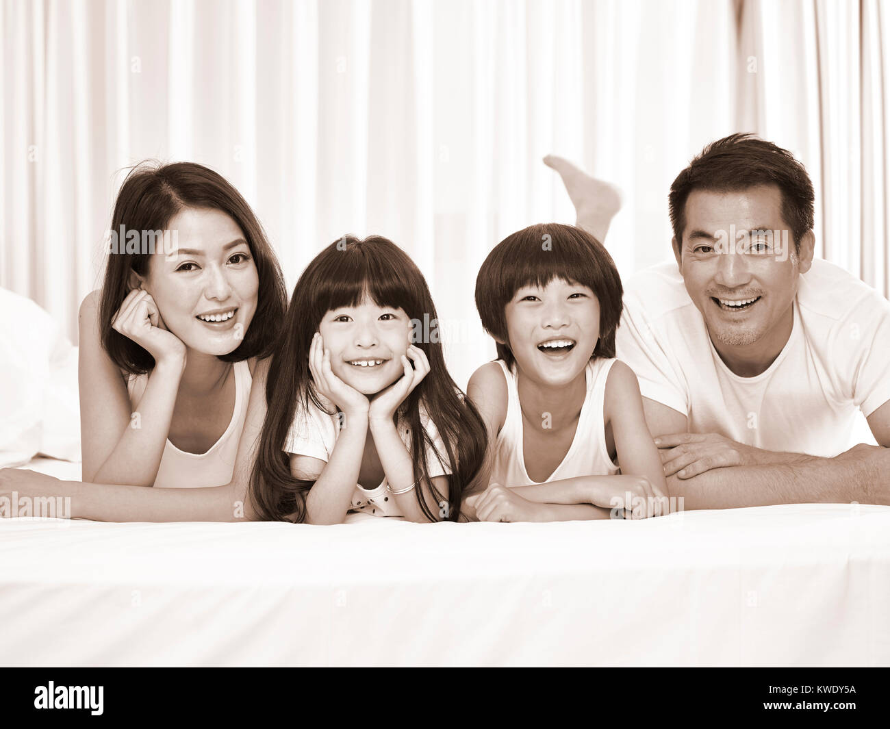 happy asian family with two children lying on front in bed looking at camera smiling, black and white sepia. Stock Photo