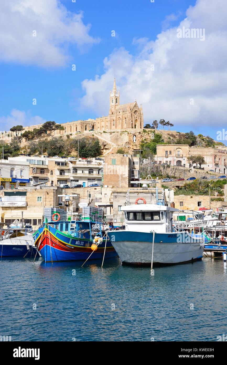 Traditional fishing boats in the harbour with the Our Lady of Lourdes church on the hillside to the rear, Mgarr, Gozo, Malta, Europe. Stock Photo