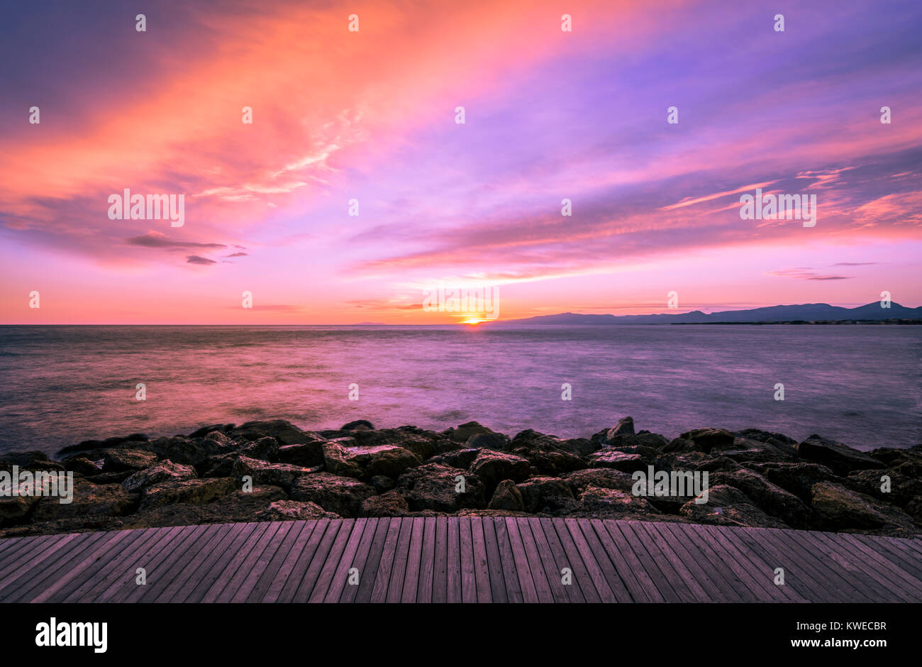 Colorful ultra violet dark sunset over the sea and with a wooden walkway and rocks on the foreground. Stock Photo
