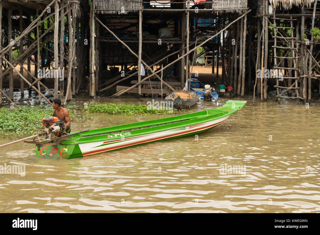 A muscular Cambodian Asian man walking in Tonle Sap Great Lake floodplain brown water, next to motor powered long thin green boat, Cambodia South East Stock Photo