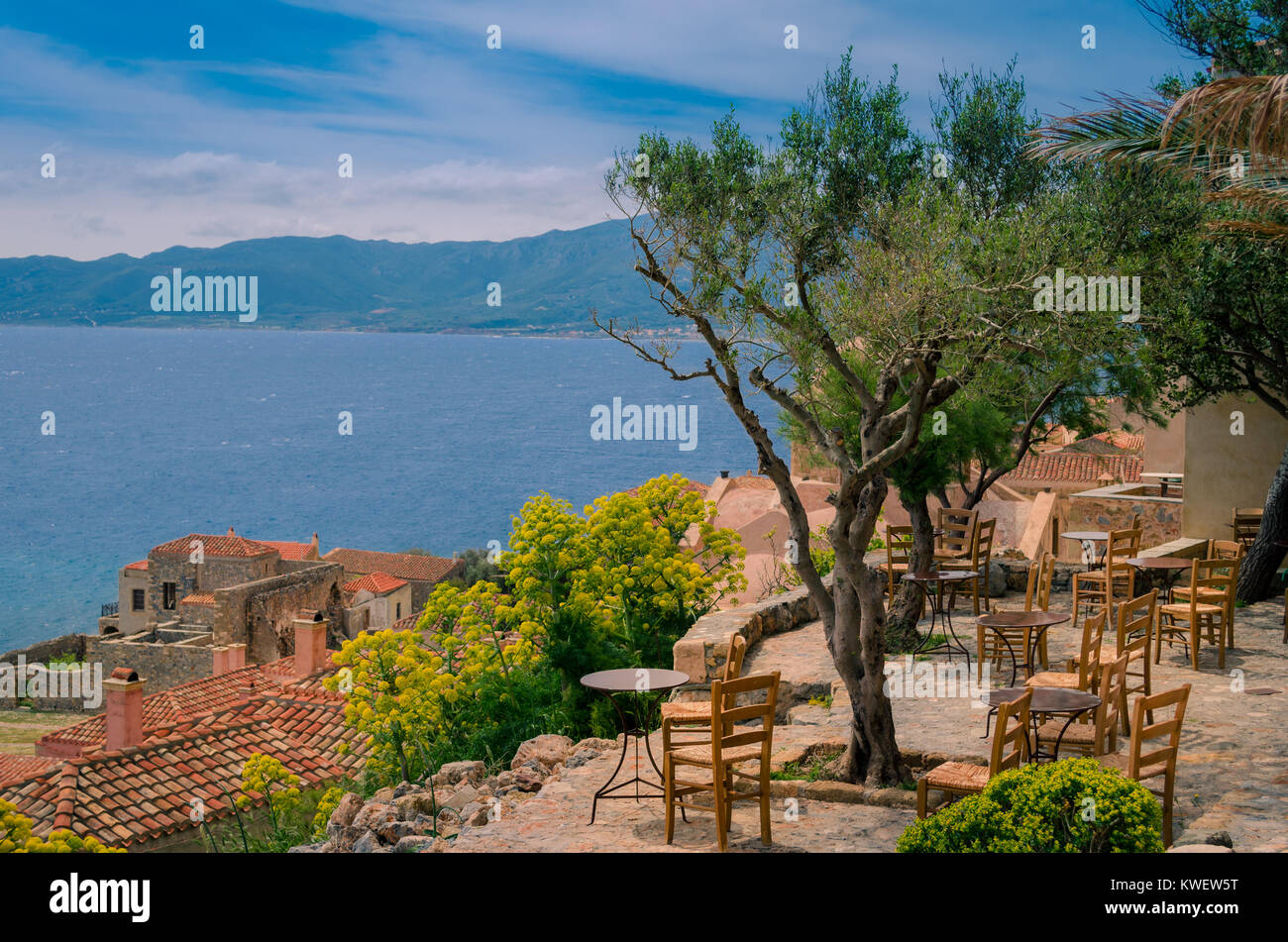 An olive tree surrounded by round iron tables and wooden chairs, typical for traditional greek cafes, overlooking the roofs of houses and the sea Stock Photo