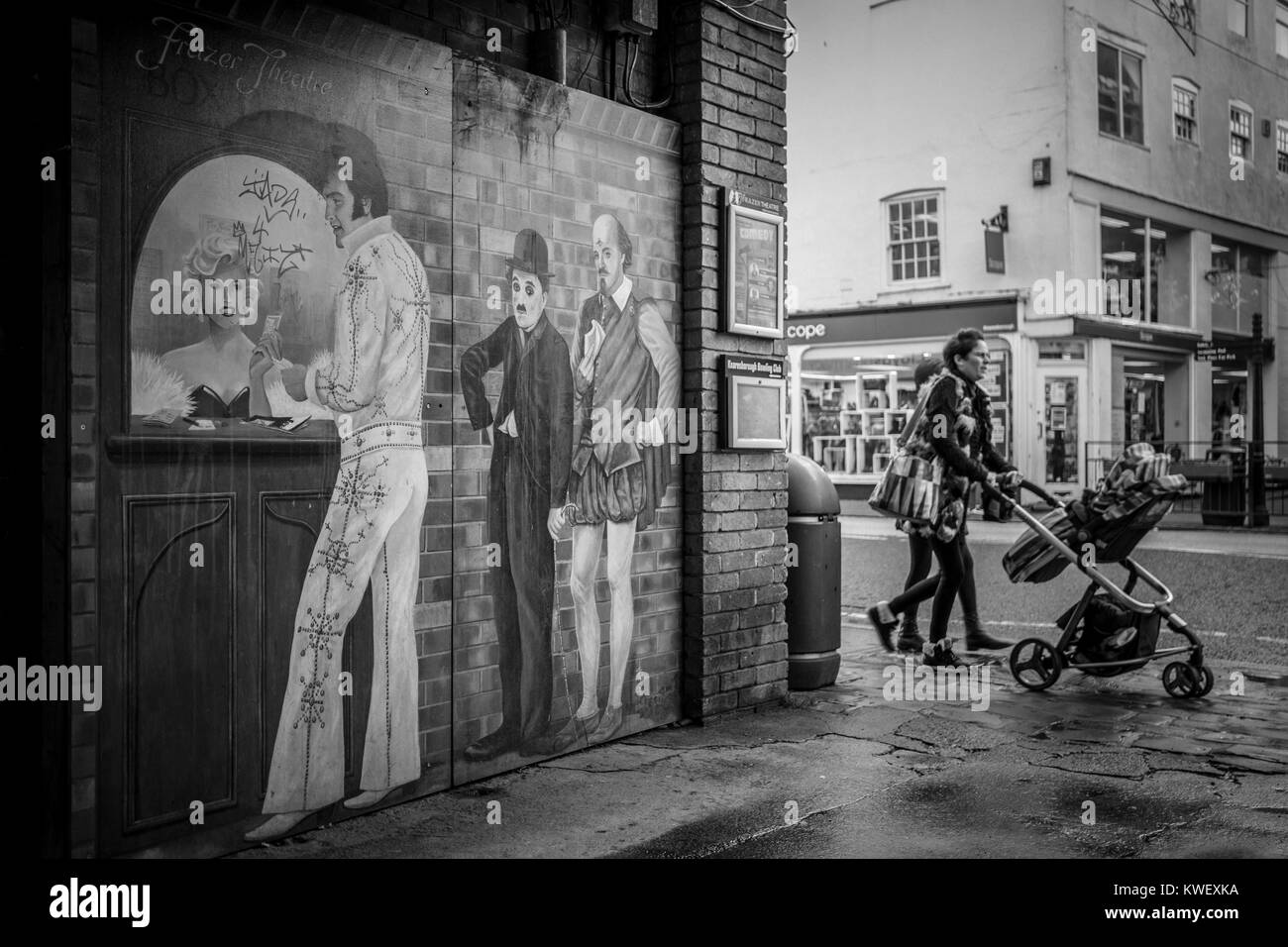 Wall mural of Elvis, Charlie Chaplin, William Shakespeare buying theatre tickets from Marilyn Monroe as woman walks past pushing a pram. Street photo. Stock Photo