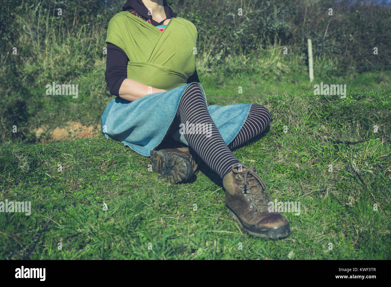 A young woman with a baby in a sling is sitting on the grass outside Stock Photo