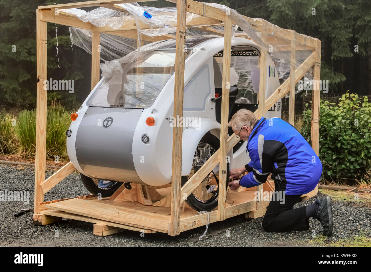 An elderly man in a blue coat kneels on the ground, using a tool to take apart a wooden shipping crate containing his new ELF electric tricycle. Stock Photo