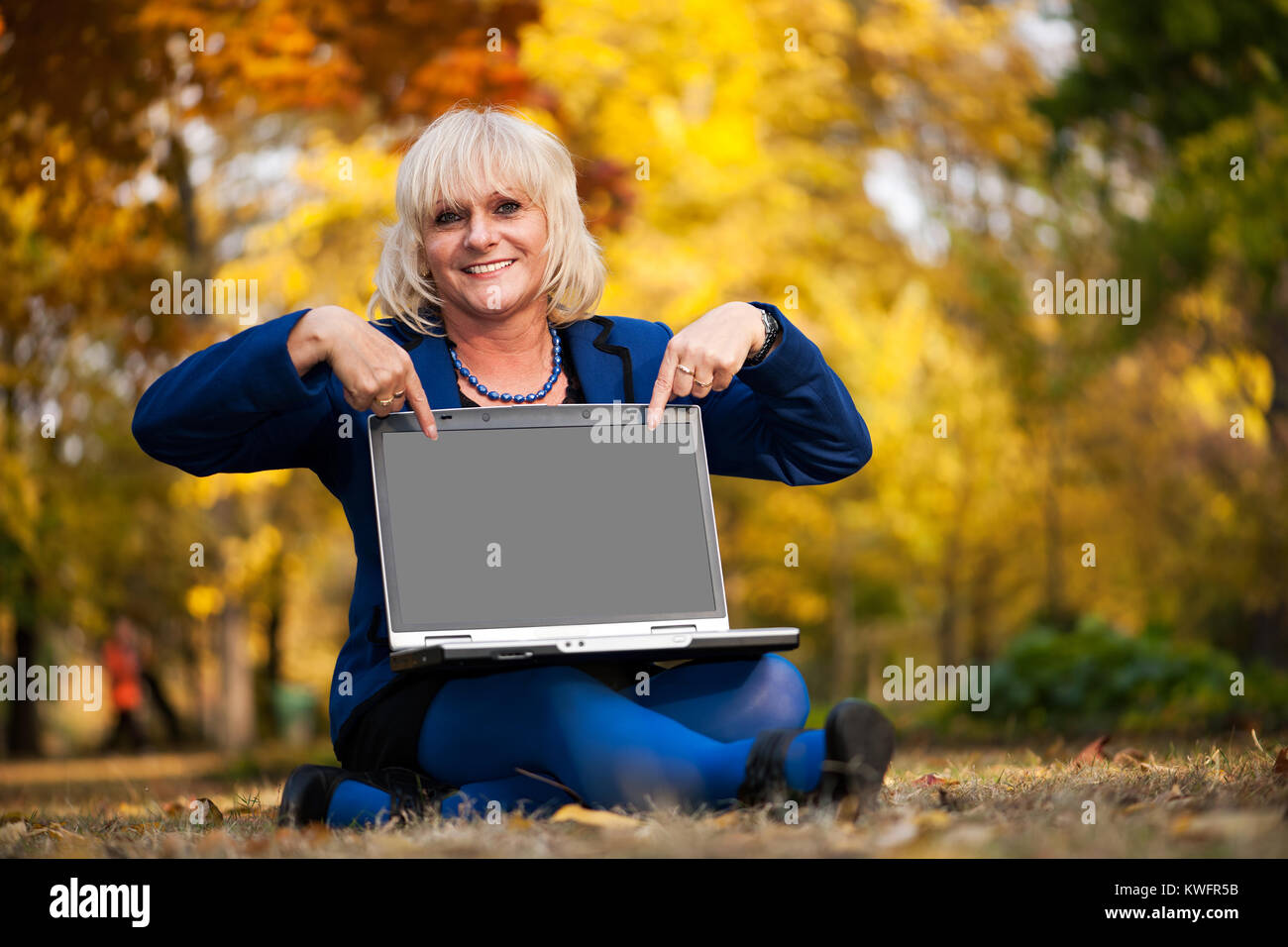 Mature woman in a beautiful fall forest holding a laptop and pointing to its empty screen Stock Photo