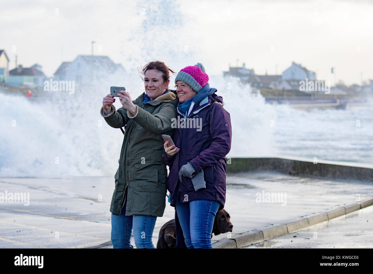 Anglesey, Wales, 3rd January 2018. UK Weather. A severe Met Office warning has been issued for the fifth storm of the UK season named Storm Eleanor. With an increase risk due the current moon state and high tides flood warnings are in place for many areas along with gales force winds likely to cause harm. Huge waves and winds crash into Trearddur Bay on Anglesey in North Wales but still time for a selfie as waves crash behind these friends on the seafront © DGDImages/Alamy Live News Stock Photo