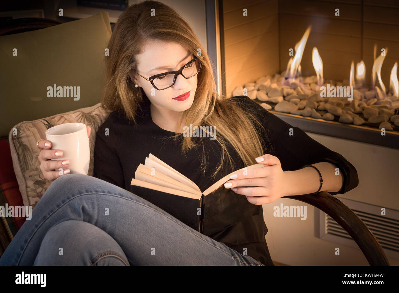 Portrait of a blonde young woman wearing eyeglasses, reading a book, sitting on a rocking chair beside a fireplace and having coffee. Stock Photo