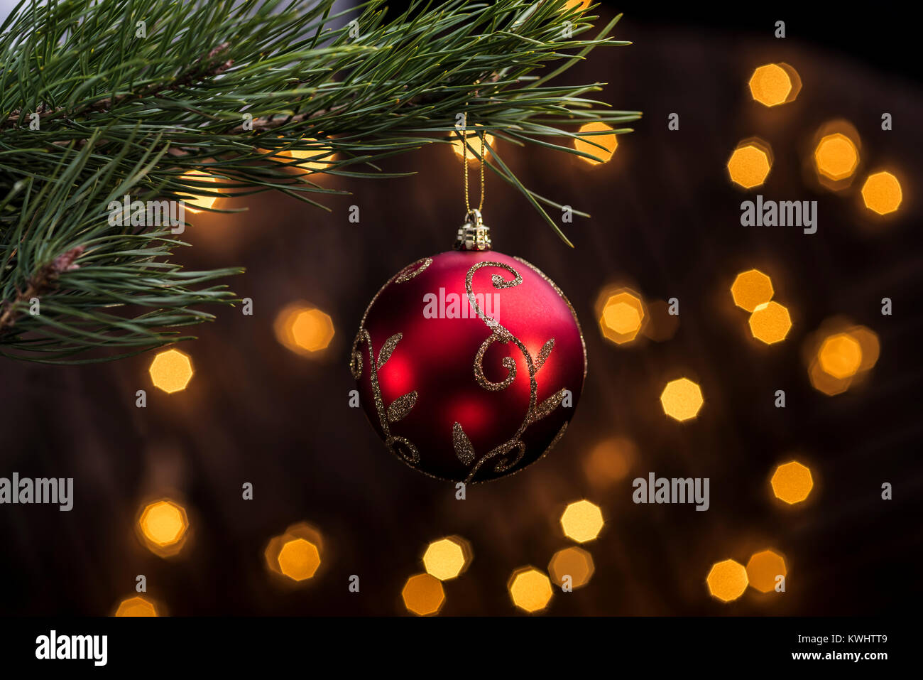 Red bauble hanging from christmas tree with festive lights out of fucus in background. Stock Photo