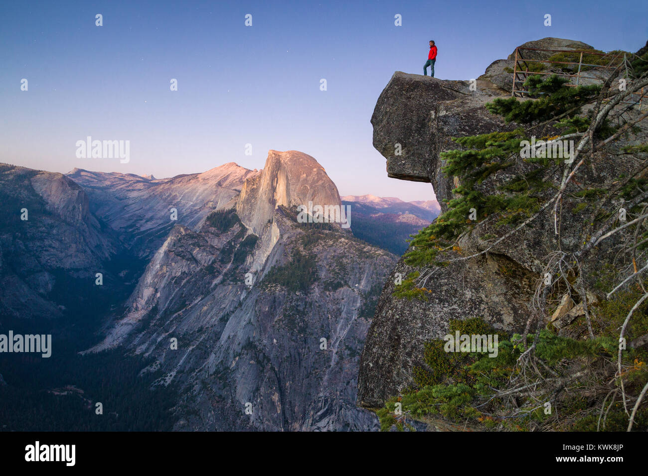 A fearless hiker is standing on an overhanging rock looking towards famous Half Dome at Glacier Point at sunset, Yosemite National Park, California Stock Photo