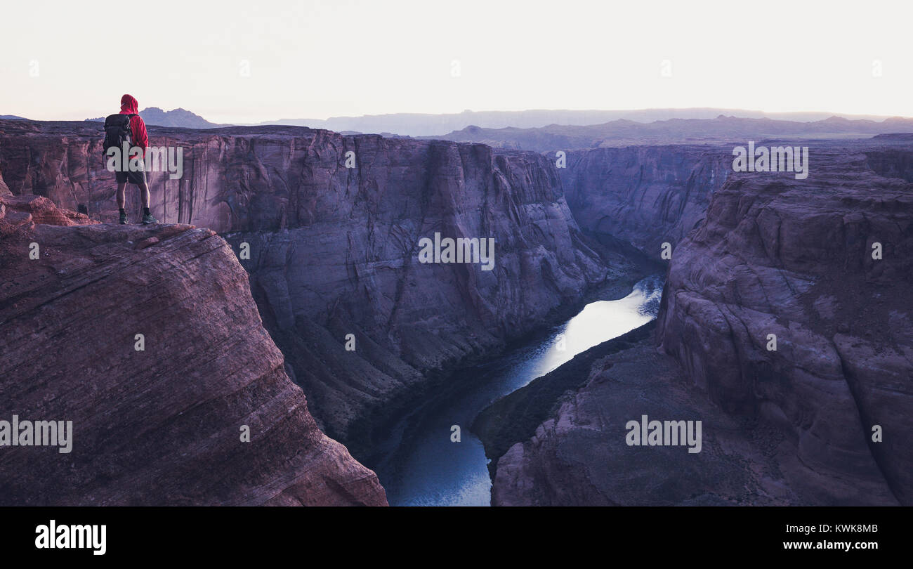 A male hiker is standing on steep cliffs enjoying the beautiful view of Colorado river flowing at famous Horseshoe Bend overlook in mystic post sunset Stock Photo