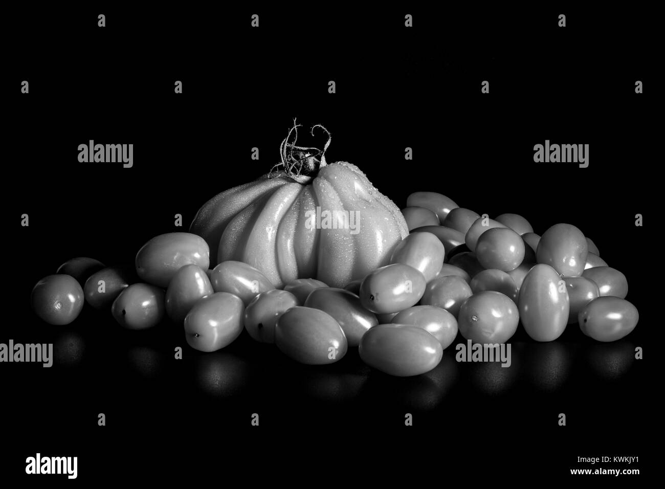 A black and white image of large beefsteak tomato surrounded by small baby plum tomatoes. Lighting emphasises shape and texture. Stock Photo