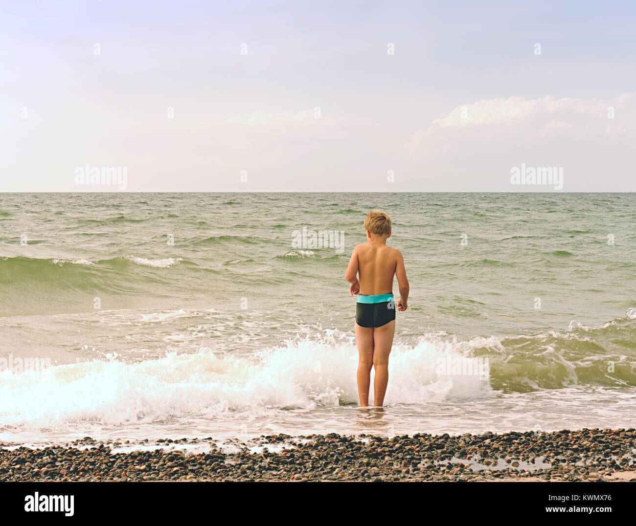 Cute fair-haired blond boy walking on beach into foamy sea waves.  Windy summer day, cloudy  blue sky on seascape background Stock Photo