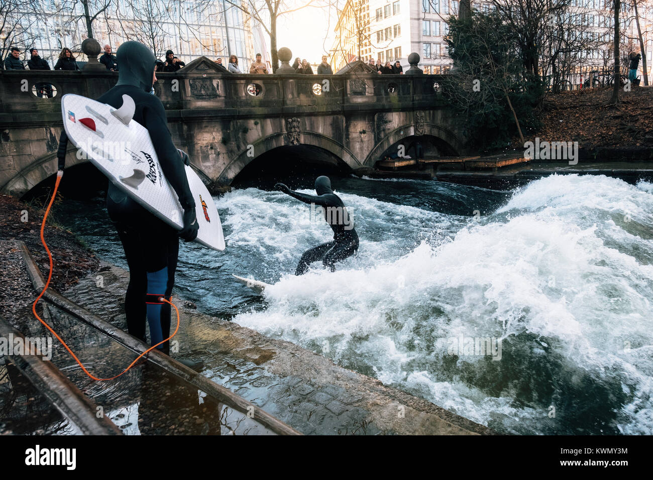 The surfer in diving suit wait their turn to ride the artificial wave on the Eisbach, dangerous water Isar river channel at Englischer Garden. Surfing Stock Photo