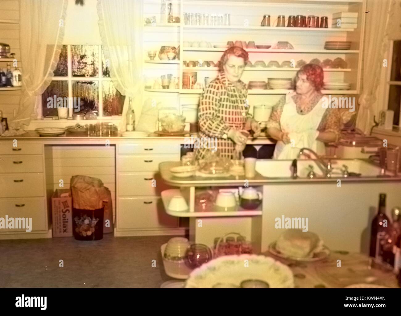 Two mature women cook in a suburban kitchen, with Soilax laundry detergent box visible, Eureka, California, 1950. Note: Image has been digitally colorized using a modern process. Colors may not be period-accurate. Stock Photo