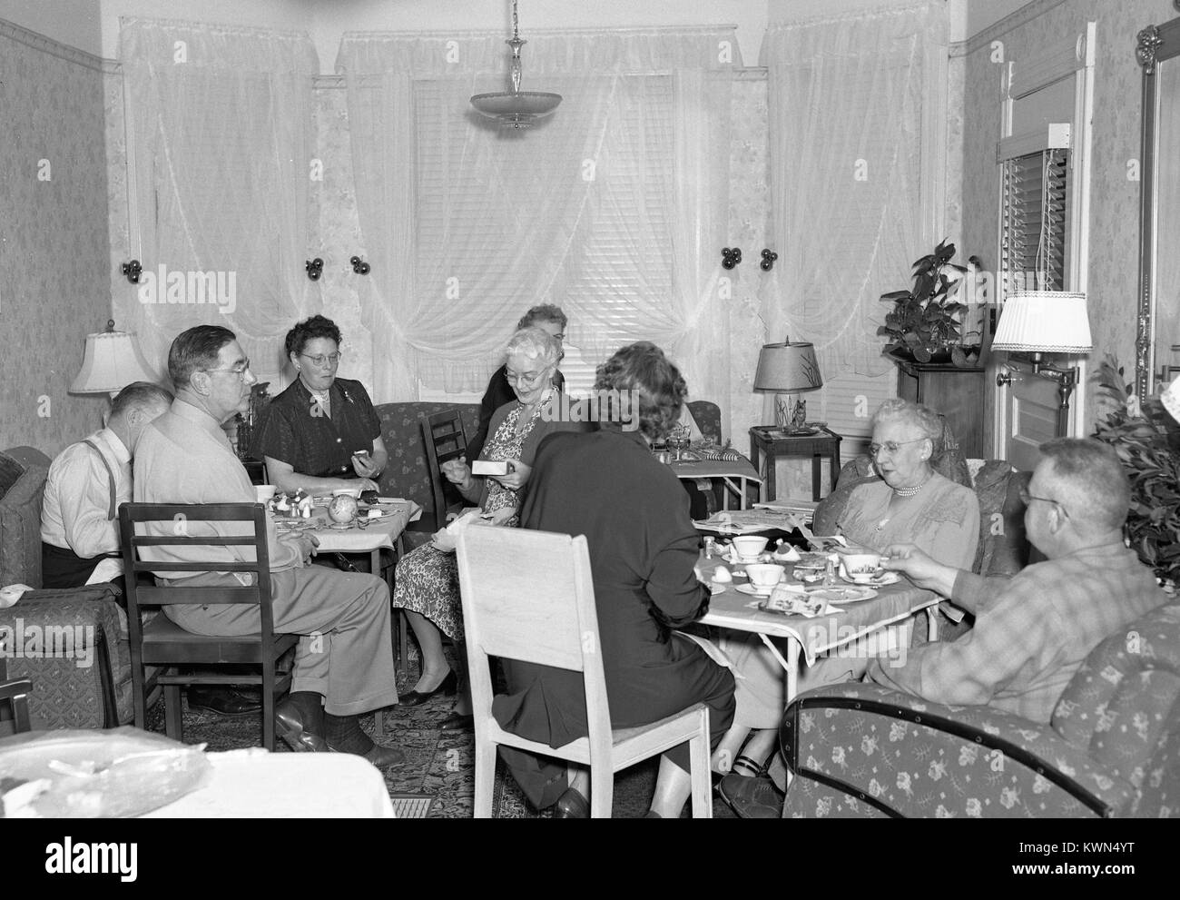A large group of mature adults sits at various tables in a suburban home and converses over tea, Eureka, California, 1950. Stock Photo