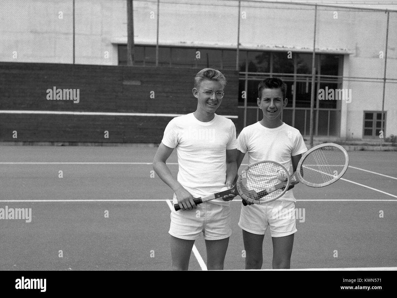 Two young boys wearing white athletic apparel hold tennis rackets and post for a photograph on a tennis court prior to a match, Monterey, California, 1950. Stock Photo