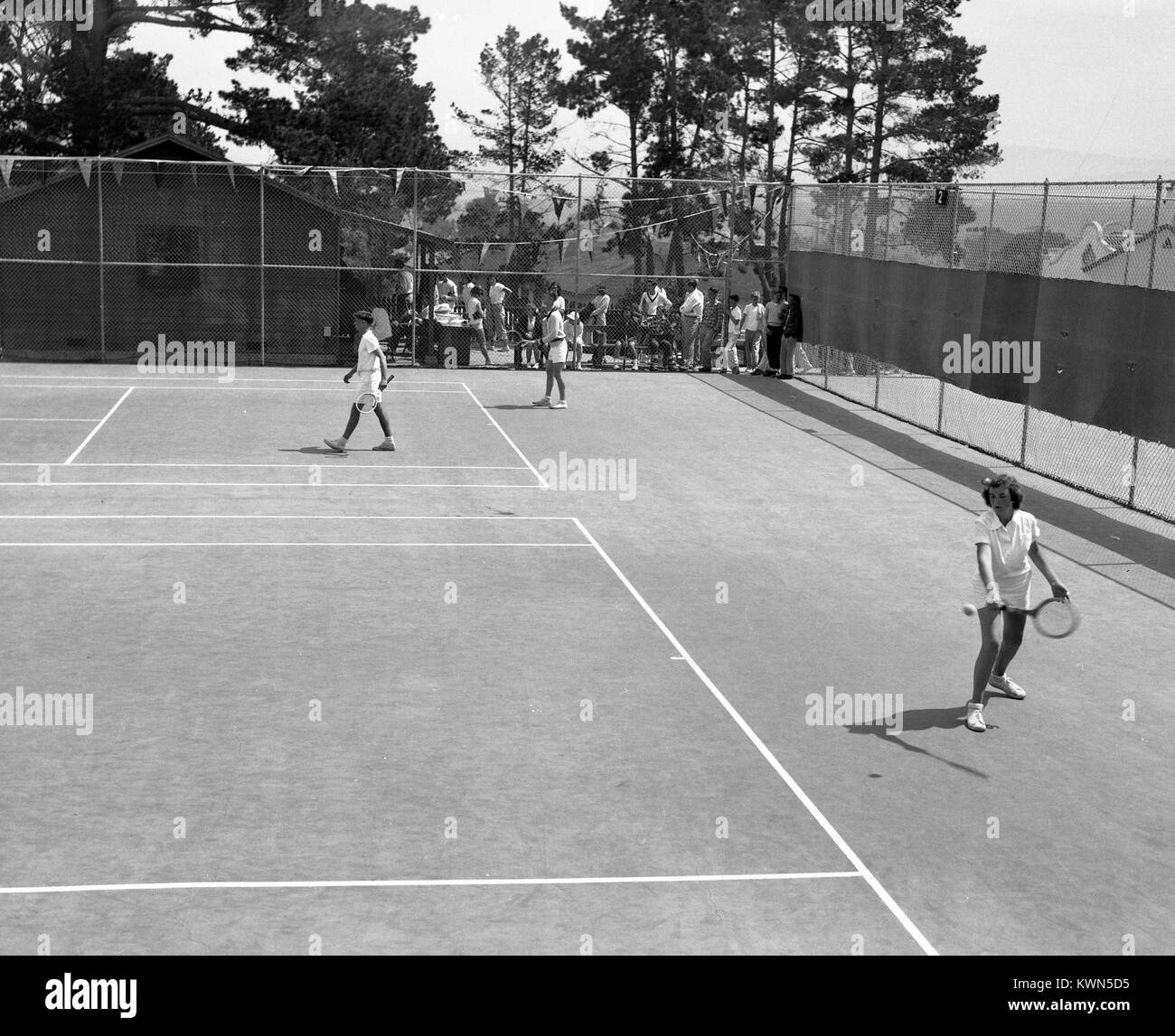 A young woman hits a tennis ball while several young men play tennis on a court behind her, Monterey, California, 1950. Stock Photo