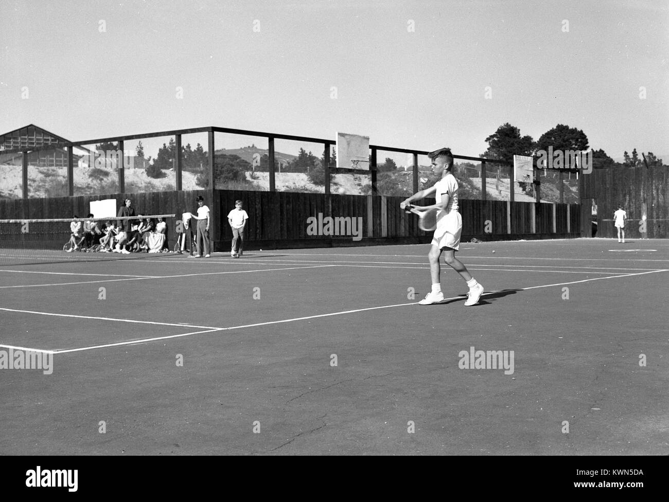 A young boy lines up a shot while playing tennis, as a group of his peers look on, Monterey, California, 1950. Stock Photo
