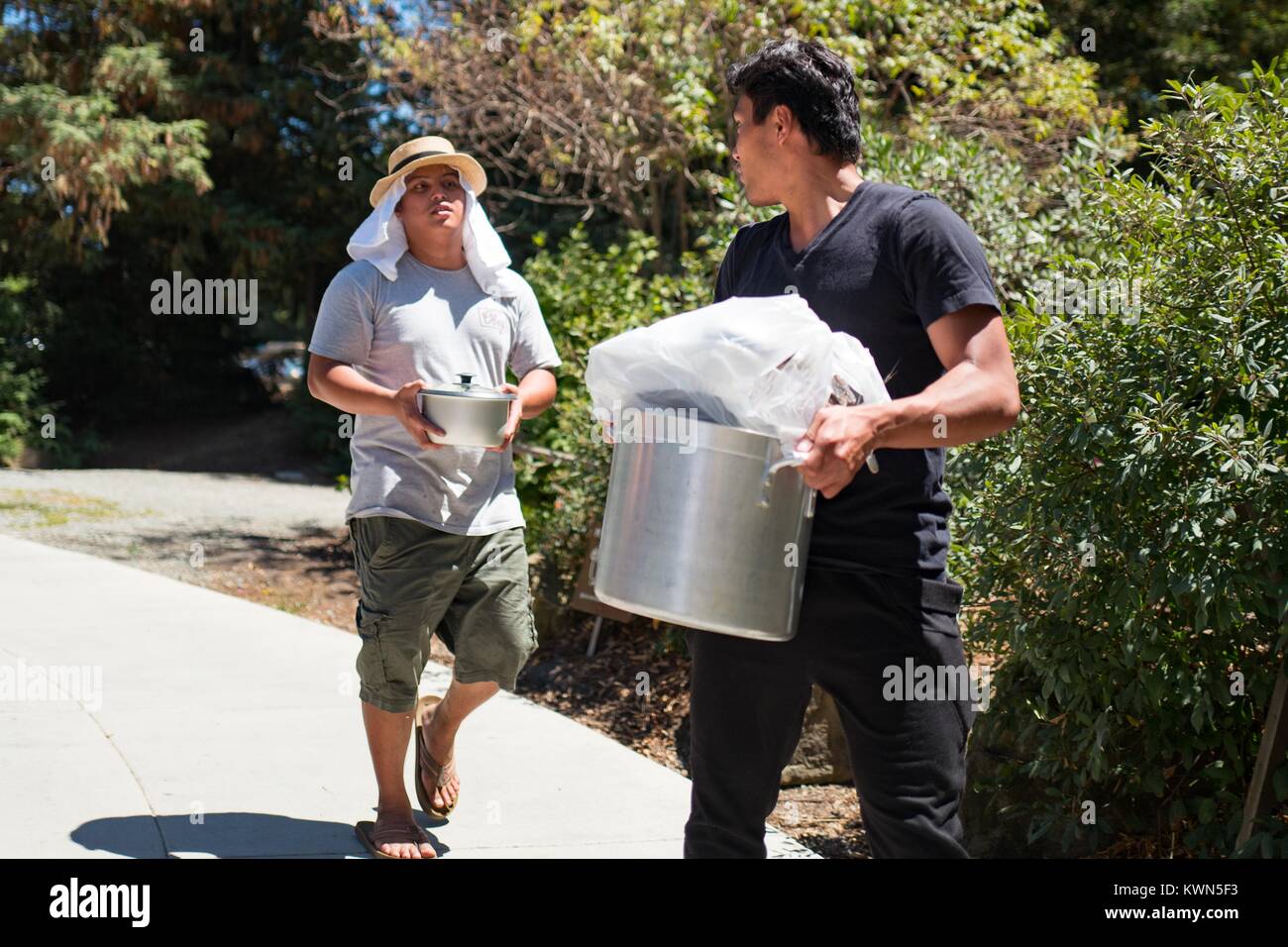 Two men, one wearing a straw hat and head scarf to protect himself from the hot sun, carry large metal pots of food towards a picnic site at Lake Chabot Regional Park, an East Bay Regional Park in the San Francisco Bay Area town of San Leandro, California, July 16, 2017. Stock Photo