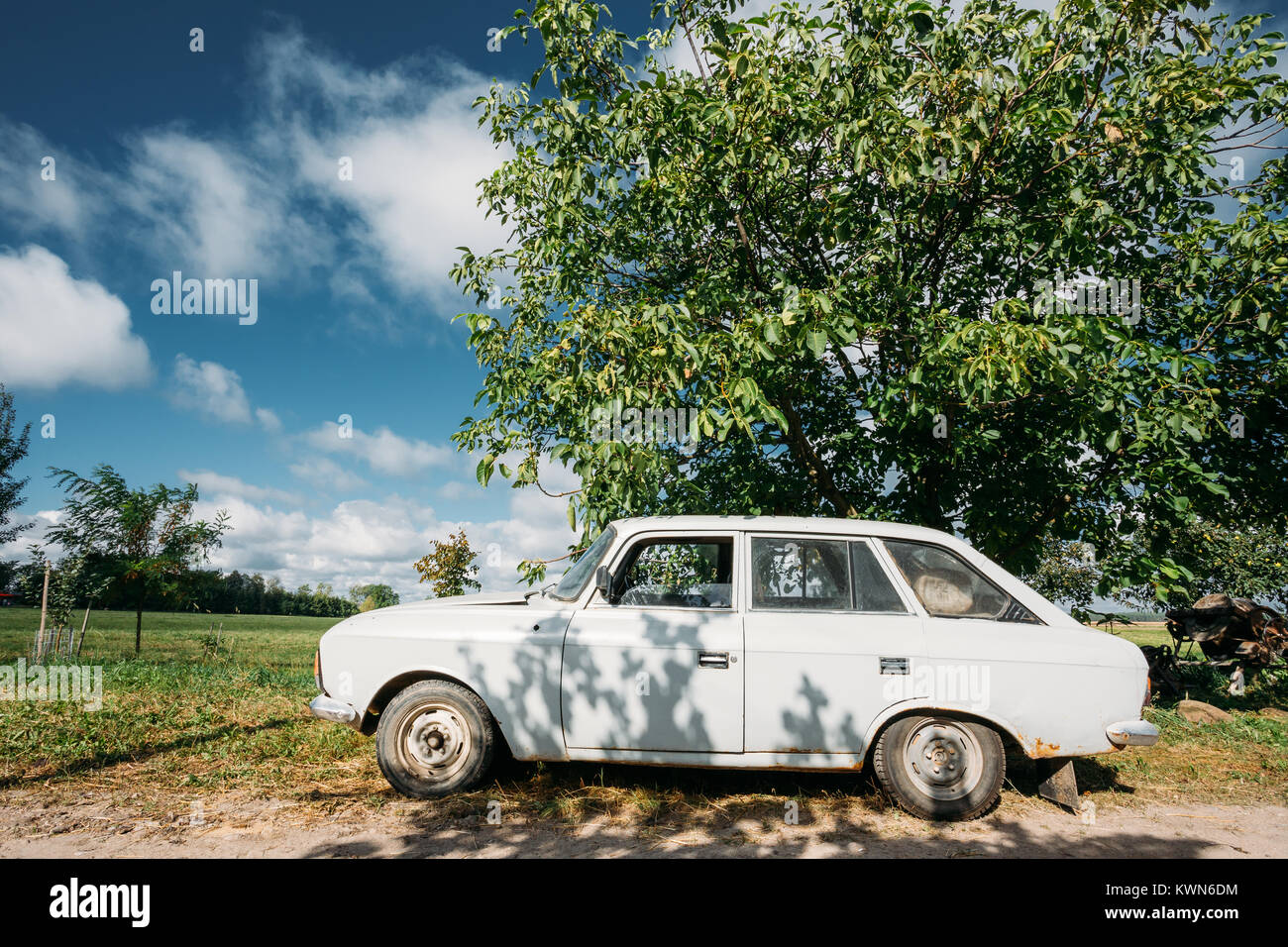 Old White Izh 21251 Kombi Parking On A Roadside Of Country Road On A Background Of Green Tree And Meadow In Sunny Day With Blue Sky. Stock Photo