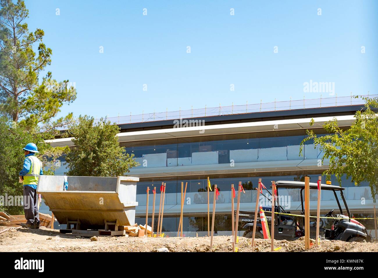 A construction worker in a high visibility vest and blue hard hat stands with construction equipment in front of a portion of the Apple Park, known colloquially as 'The Spaceship', the new headquarters of Apple Inc in the Silicon Valley town of Cupertino, California, July 25, 2017. Stock Photo