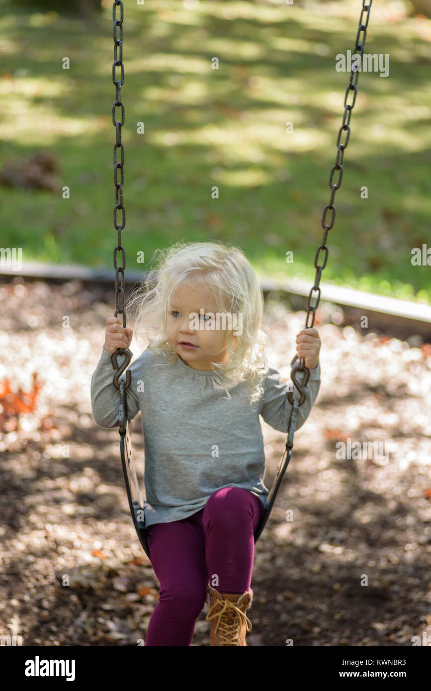 little blonde girl playing on swing set in park on a sunny autumn day Stock Photo