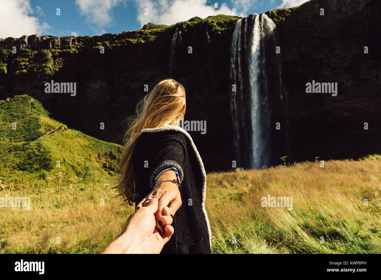 cropped shot of girl holding hand of friend and going to majestic waterfall in Iceland Stock Photo