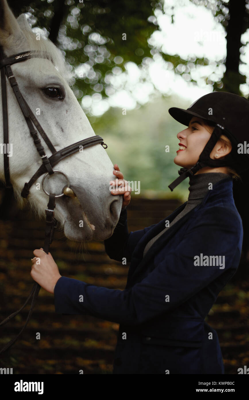 Beautiful horsewoman in blue jacket strokes white horse. Stock Photo