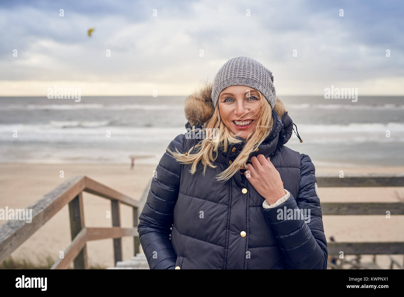 Middle-aged woman braving a cold winter day at the seaside standing on a wooden deck overlooking the beach on a breezy day smiling happily at the came Stock Photo