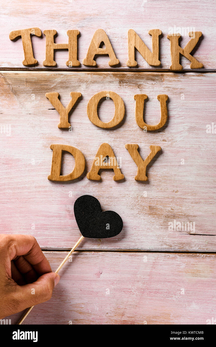 some wooden letters forming the text thank you day on a rustic wooden surface and a heart attached to a stick in the hand of a young caucasian man Stock Photo