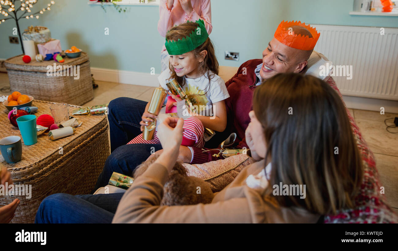 Cheerful family are relaxing at home at christmas. They are opening christmas crackers while curled up on the sofa together. Stock Photo