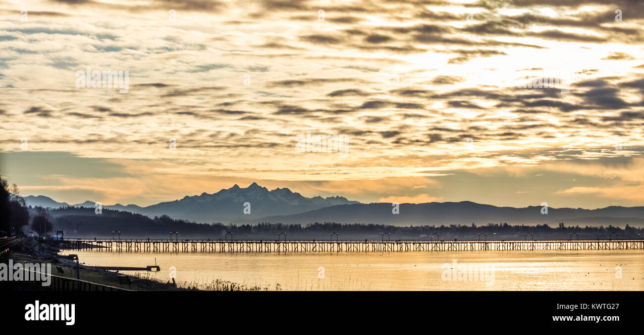 Wooden pier at White Rock extends into Semiahmoon Bay shared by Canada and USA. Pier is silhouetted below majestic, snow-capped Three Fingers Mountain Stock Photo