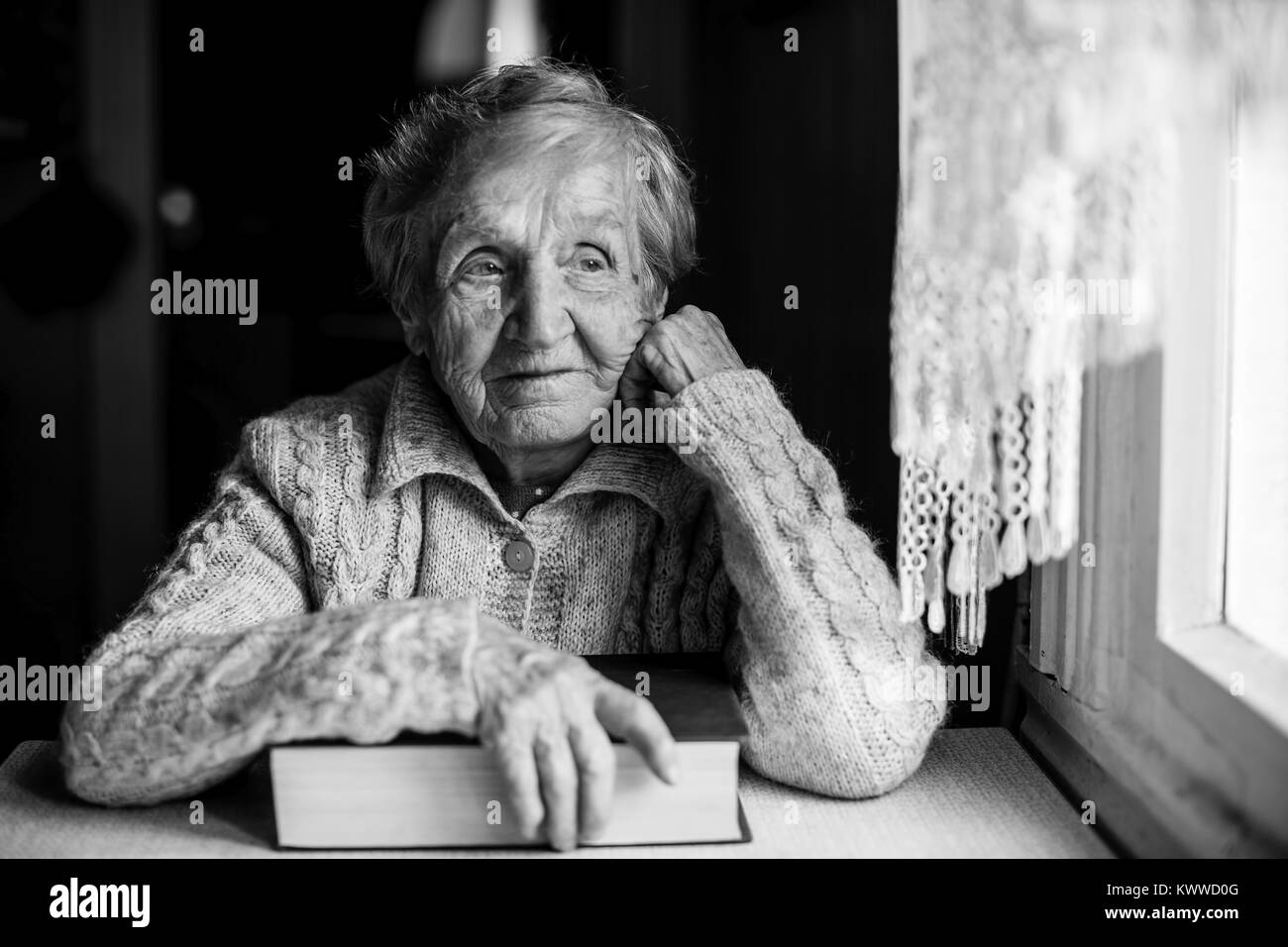 An elderly woman with a book in hand sitting at a table near the window. Stock Photo