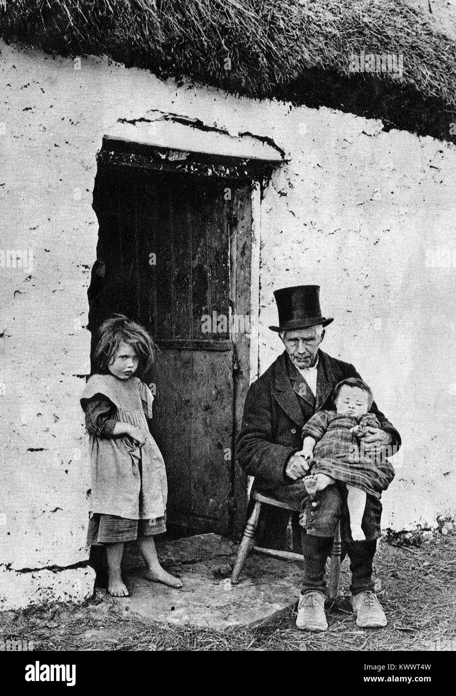 An Irishman wearing a top hat with children sitting at his thatched cottage door in 1920 Stock Photo