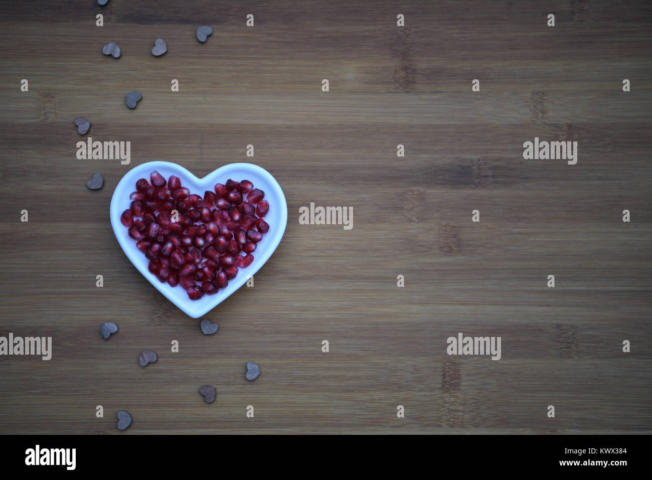 healthy food photography of red shiny juicy shiny pomegranate seeds in white heart shape dish with love heart decorations on wood background and space Stock Photo