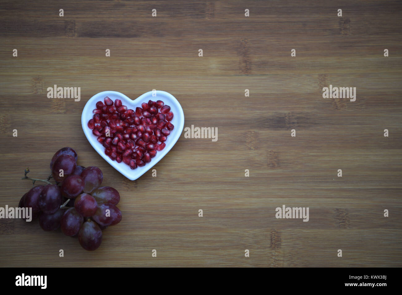 healthy food photography of red juicy shiny pomegranate seeds in love heart shape dish with a bunch of purple grapes on wood background and space Stock Photo