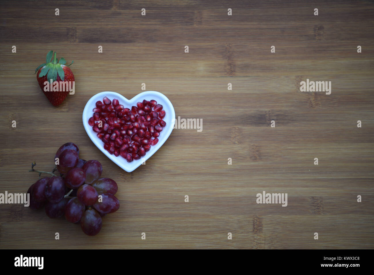 healthy food photography of red juicy shiny pomegranate seeds in love heart shape dish with a bunch of purple grapes on wood background and space Stock Photo