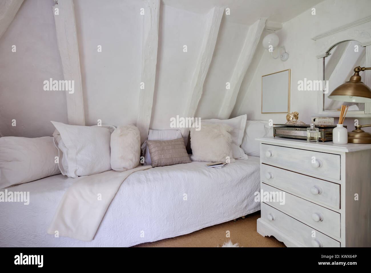 Small cottage bedroom beutifully decorated in shades of white with quilt covered daybed, matching pillows, chest of drawers, mirror, perfume botttle a Stock Photo
