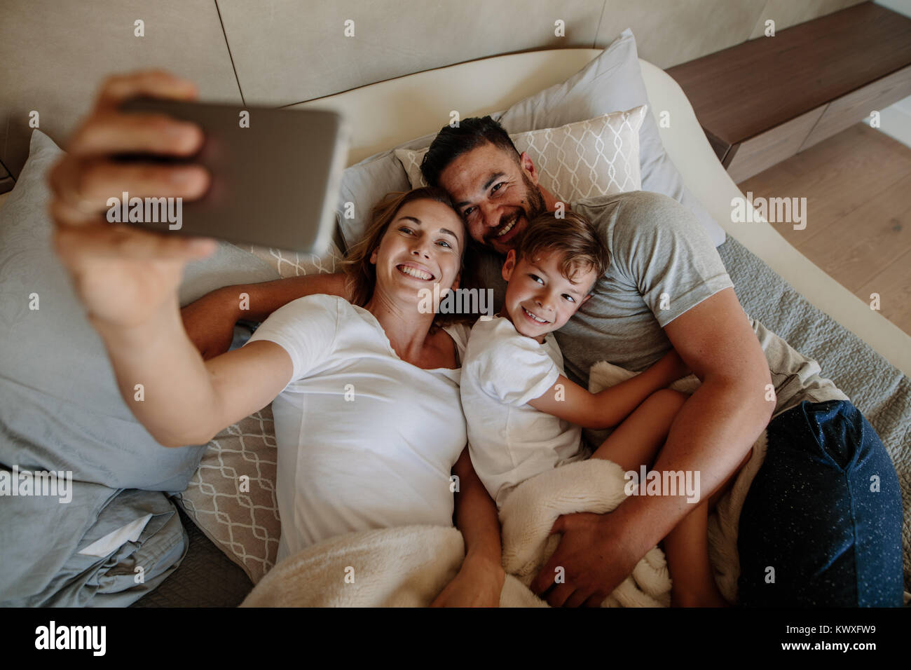 Happy young woman taking selfie with her family. Family lying on bed and taking selfie at home. Stock Photo
