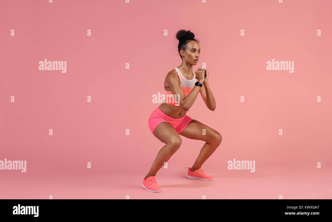 Sporty woman practicing squat exercises in studio. African woman in sportswear working out on pink background. Stock Photo