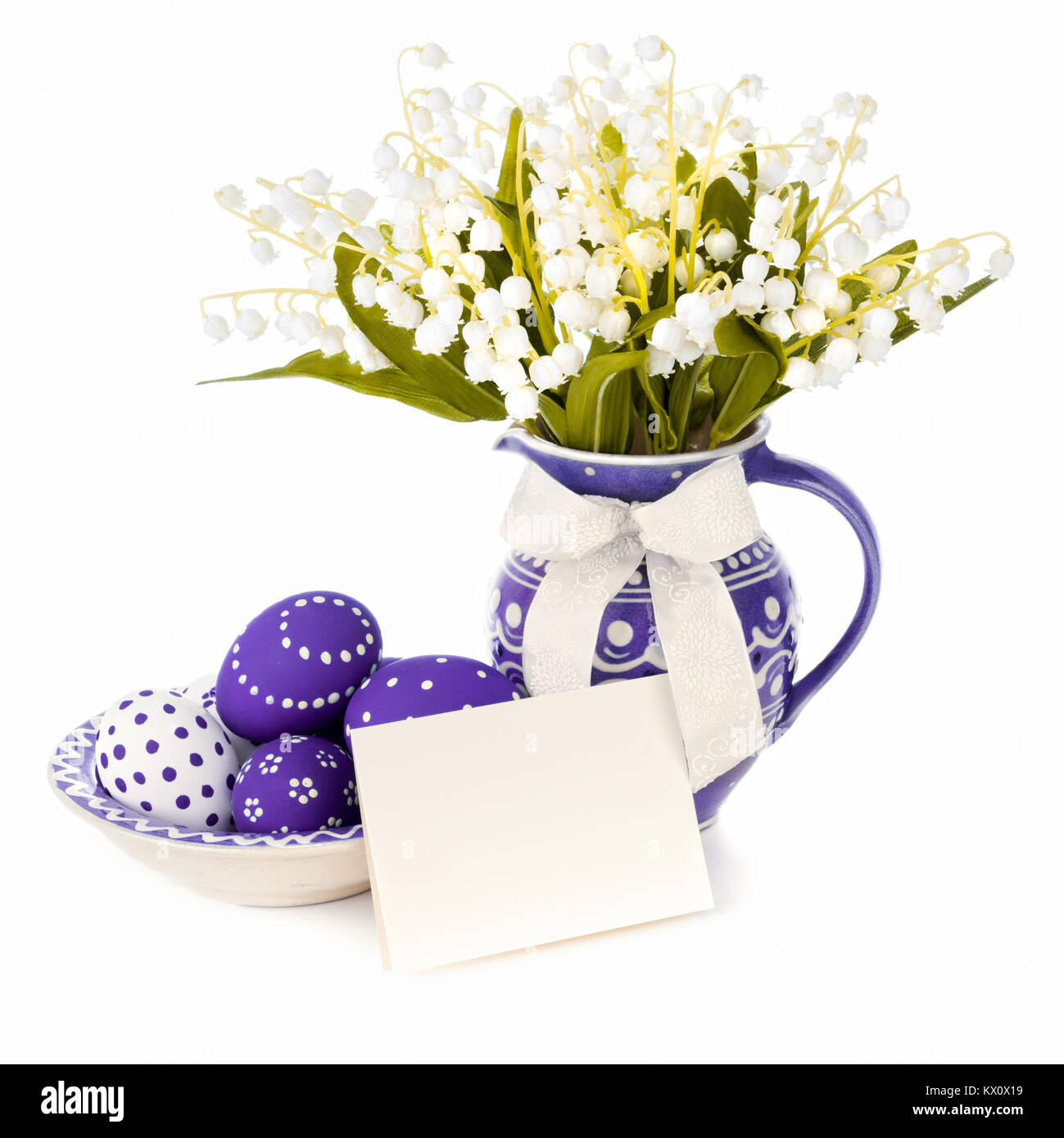 Easter eggs and artificial lily of the valley flowers in ceramic vase, isolated on white. Focus on the eggs. Space for your greeting on the blank card Stock Photo