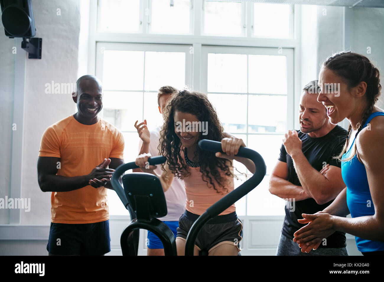 Group of people cheering on their female friend riding a stationary bike while working out together in a gym Stock Photo