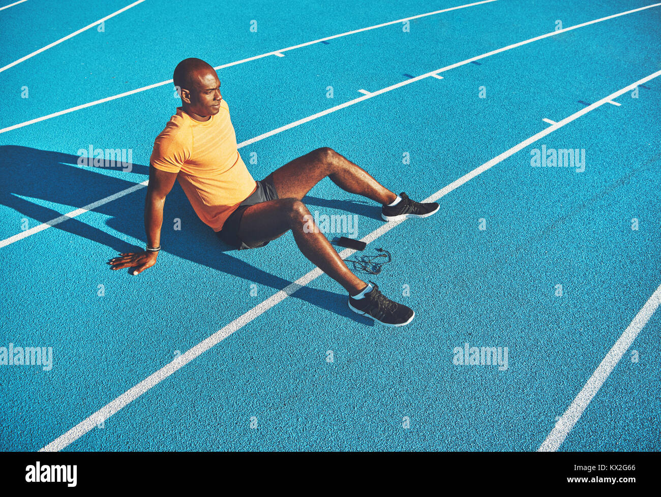 Fit young African runner sitting alone with his cellphone on a track preparing for a run on a sunny day Stock Photo