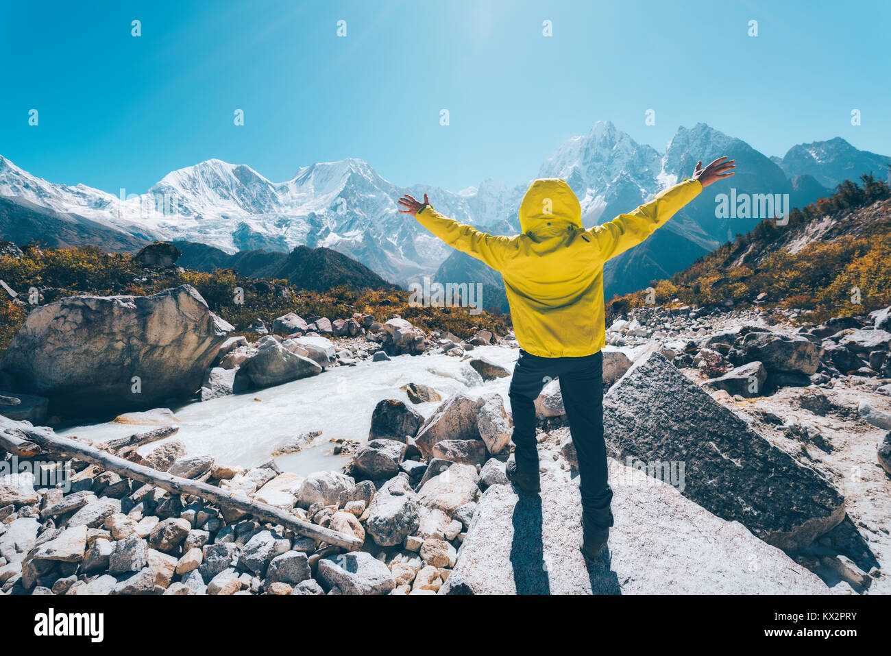 Standing man with raised up arms near the river looking on snow covered mountains in bright day. Landscape with traveler, high rocks with snowy peaks, Stock Photo