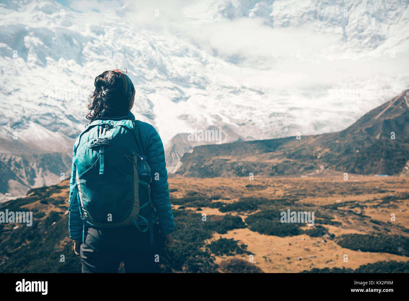 Standing woman with backpack looking on beautiful snow covered mountains in clouds at sunset. Landscape with girl, high rocks with snowy peaks, yellow Stock Photo